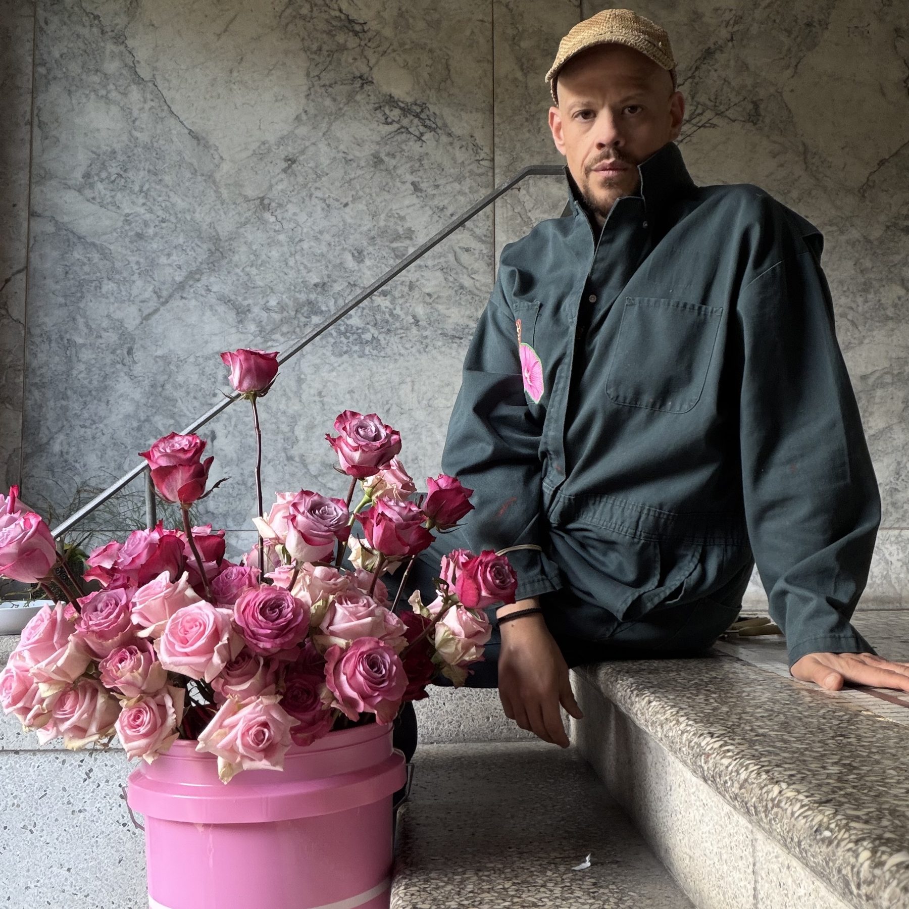 Brandon Joseph in a dark work jacket and woven cap, seated on stone steps beside a pink bucket overflowing with roses